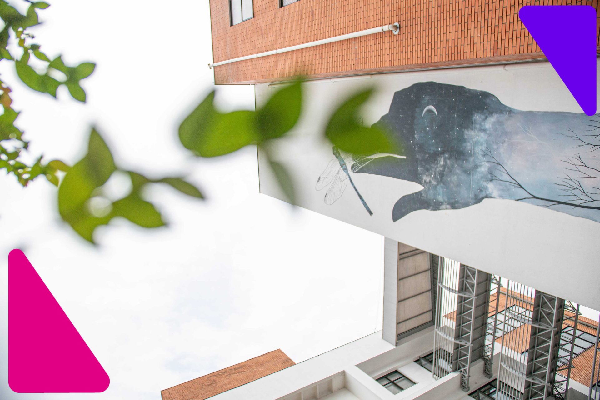 Vista inferior da fachada de um prédio de tijolos à vista na Univates, com destaque para um mural artístico pintado na estrutura e folhas verdes em primeiro plano, exibindo a infraestrutura da universidade.